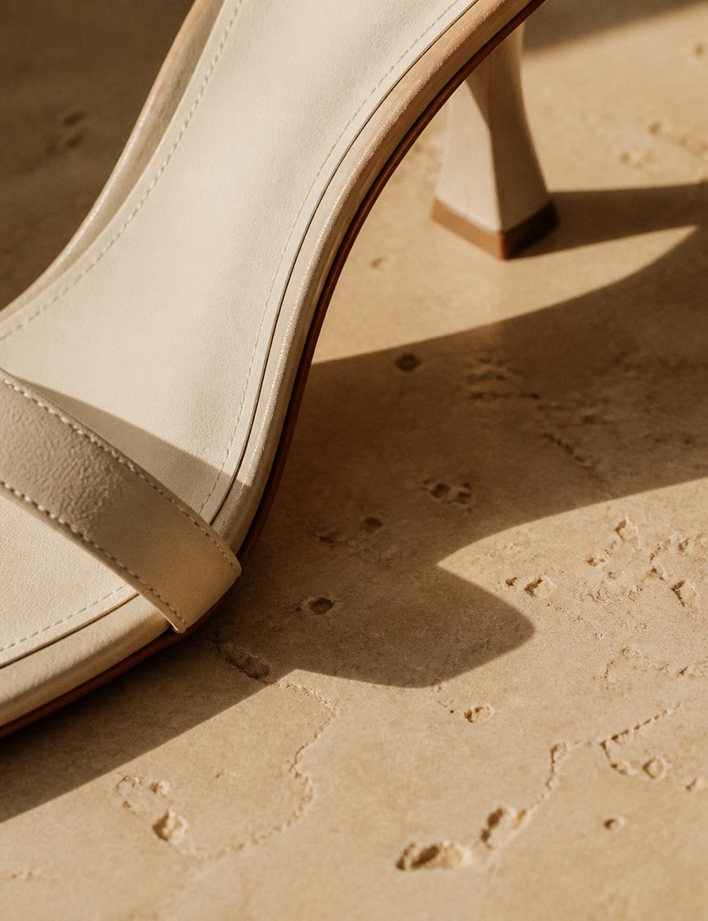 Cropped detail of a cream heeled sandal on sunlit travertine